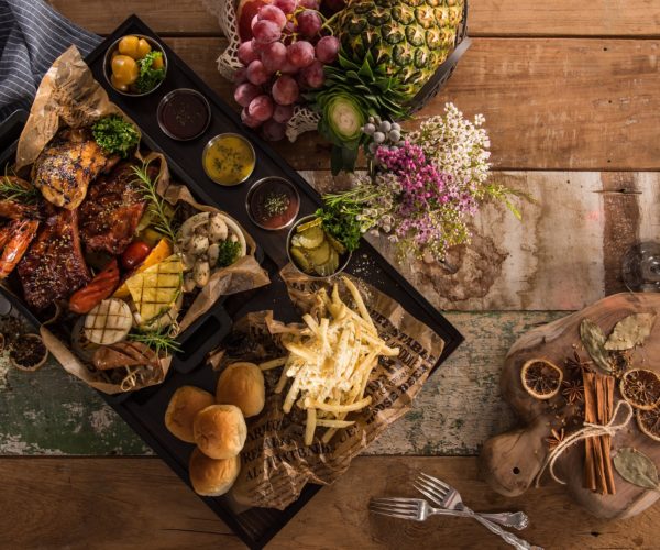 Plateau garnit avec des crevettes roties, viandes caramelisées, condiments tels que concombres, sel, poivre. Frites, ananas grillé. Une bouteille de vin rouge qui semblerait être du Médoc et un verre de vin remplit de vin au restaurant la table de 3 à Bordeaux